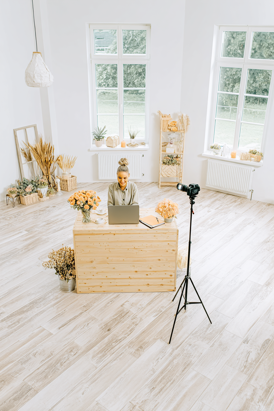 A smiling woman sits at a wooden desk with a laptop and flowers, recording content in a bright, minimal home studio with a camera setup.
