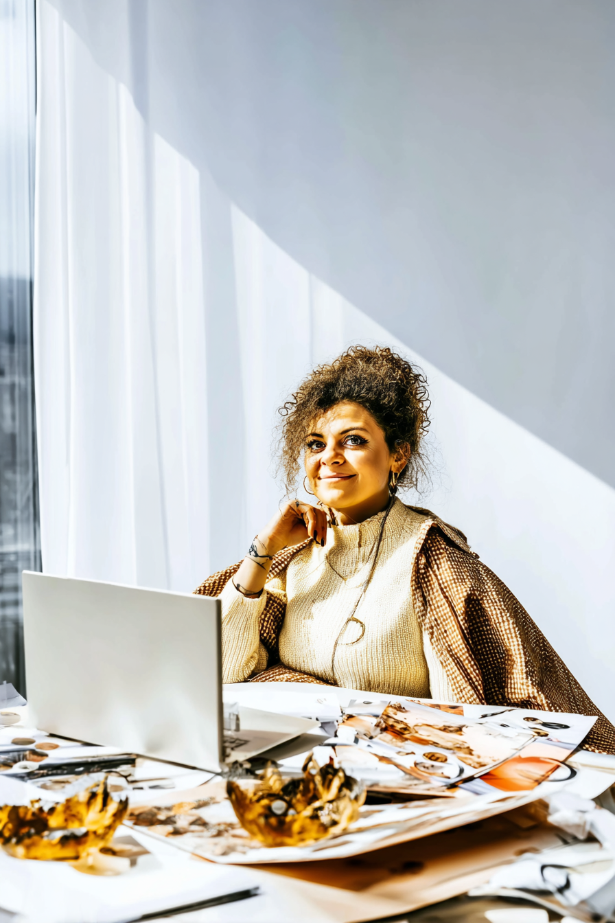 A confident woman in a cozy outfit working on a laptop surrounded by creative sketches and papers in a sunlit office.