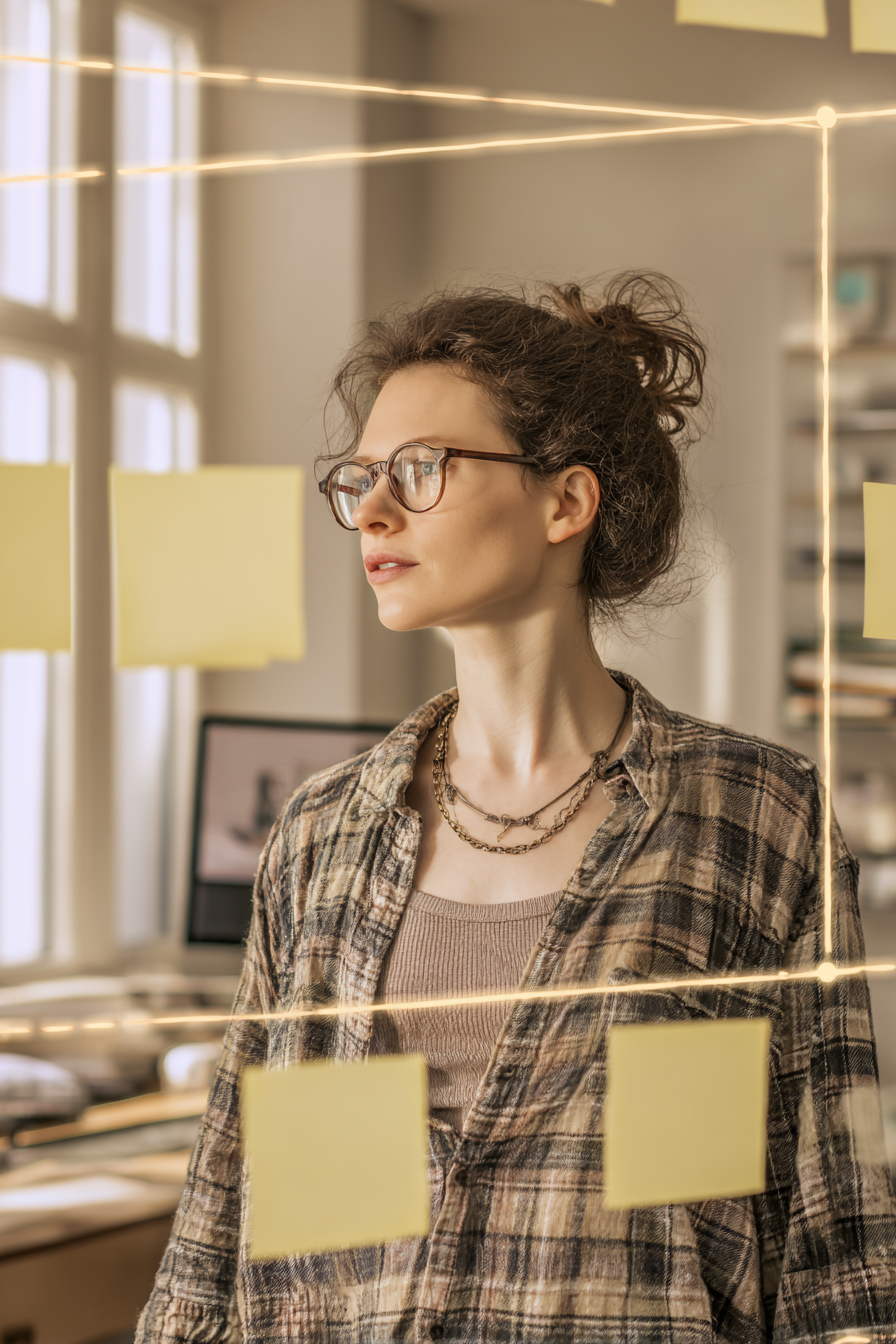 A focused woman wearing glasses stands near a glass board with sticky notes, planning tasks in a creative workspace.